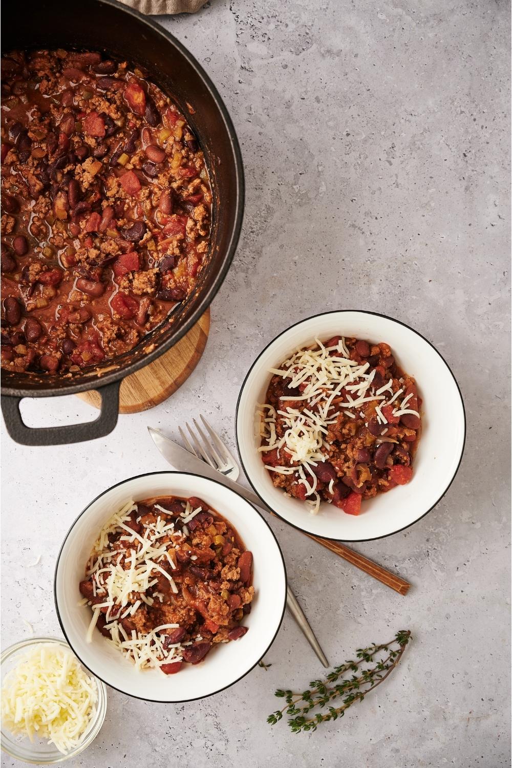 Two bowls of chili on a grey counter with a fork and knife, dried sprigs of thyme, a small bowl of cheese, and a large pot of chili resting on a wood board.