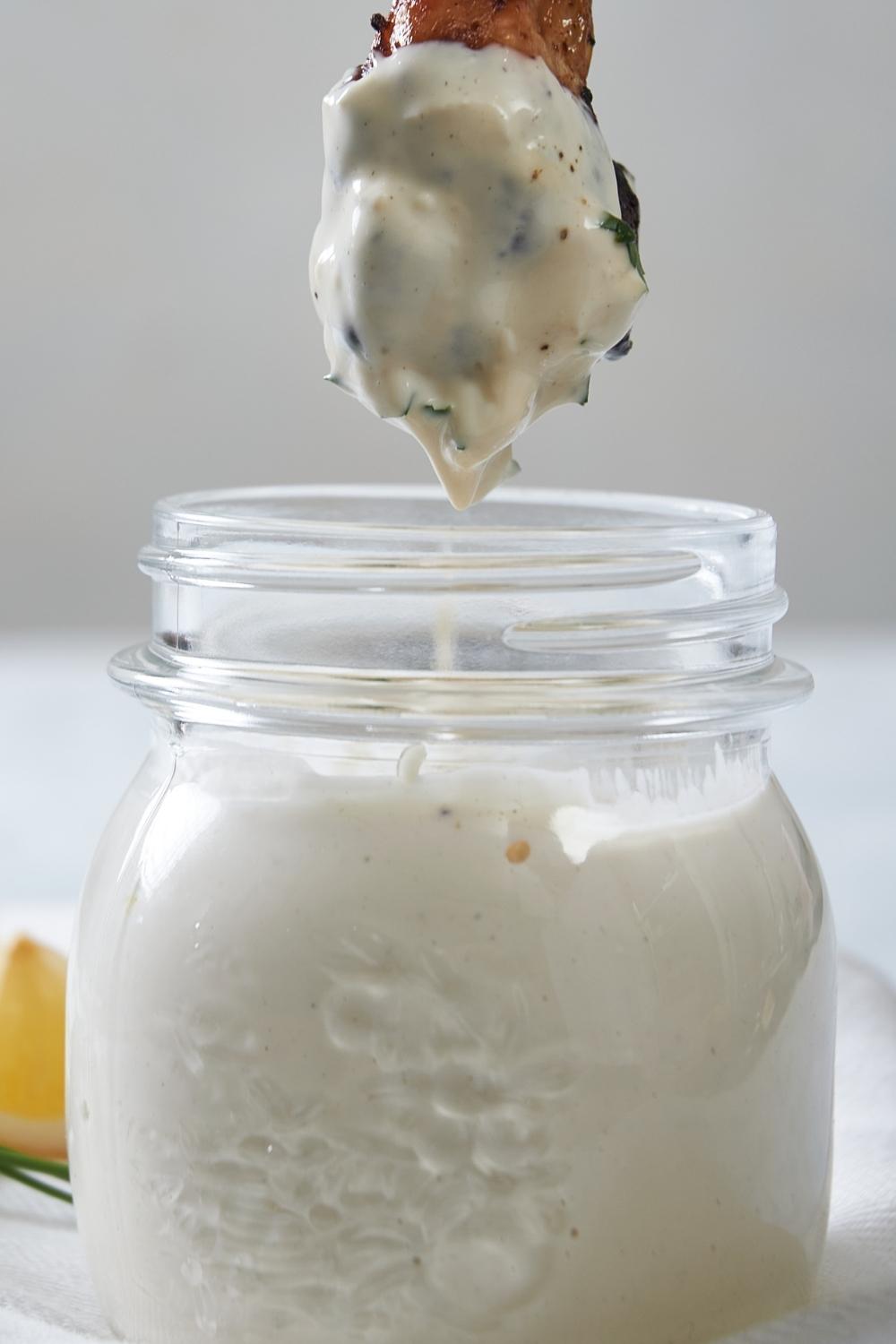 A small mason jar with homemade Alabama white sauce in it. A chicken wing has just been dipped into it and is suspended over top of the jar.