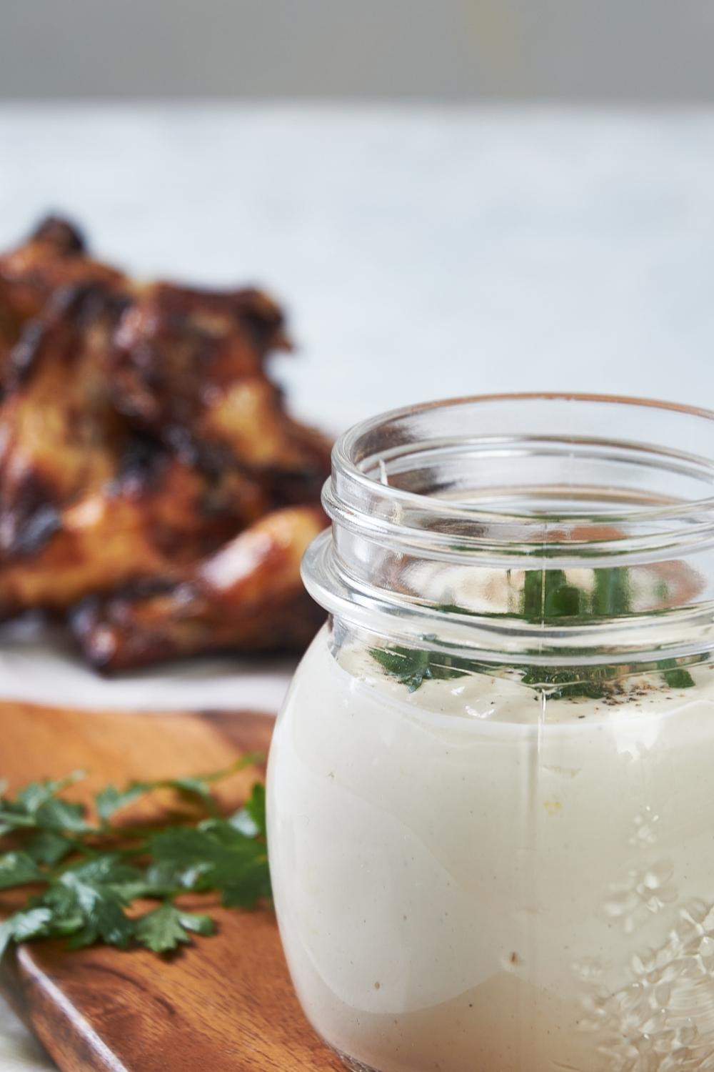 A small mason jar with homemade Alabama white sauce in it. The jar is sitting on a cutting board, and there are chicken wings on the other side of the same cutting board.