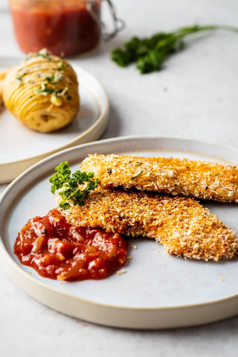 two pieces of panko chicken in a white plate with parsley for garnish and some marinara sauce on the side