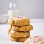 a stack of four sugar cookies, two are glazed. in the foreground is another glazed sugar cookie and in the background is a small bowl and a glass bottle of milk