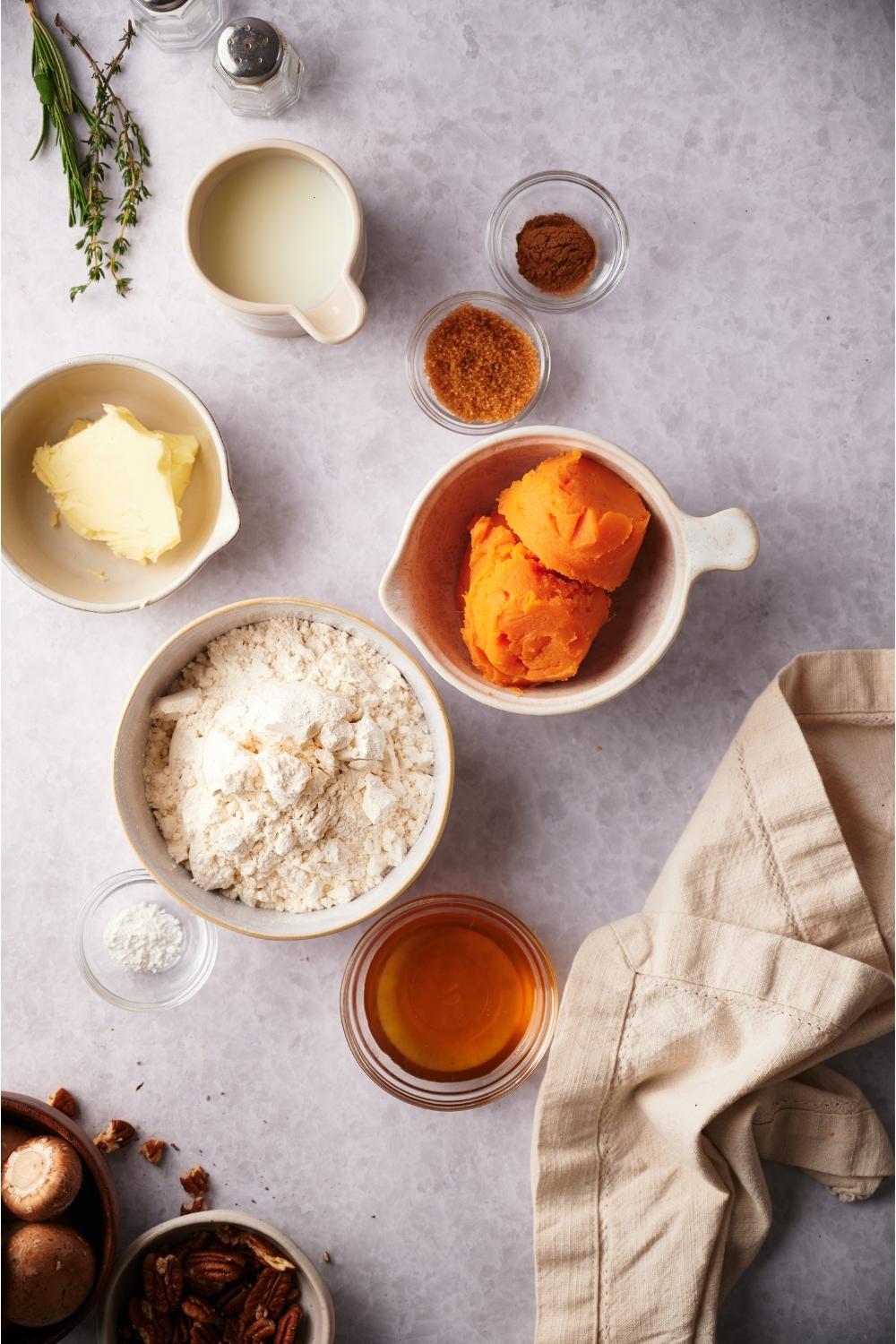 An assortment of ingredients for sweet potato biscuits including bowls of flour, sweet potatoes, milk, butter, maple syrup, and spices.