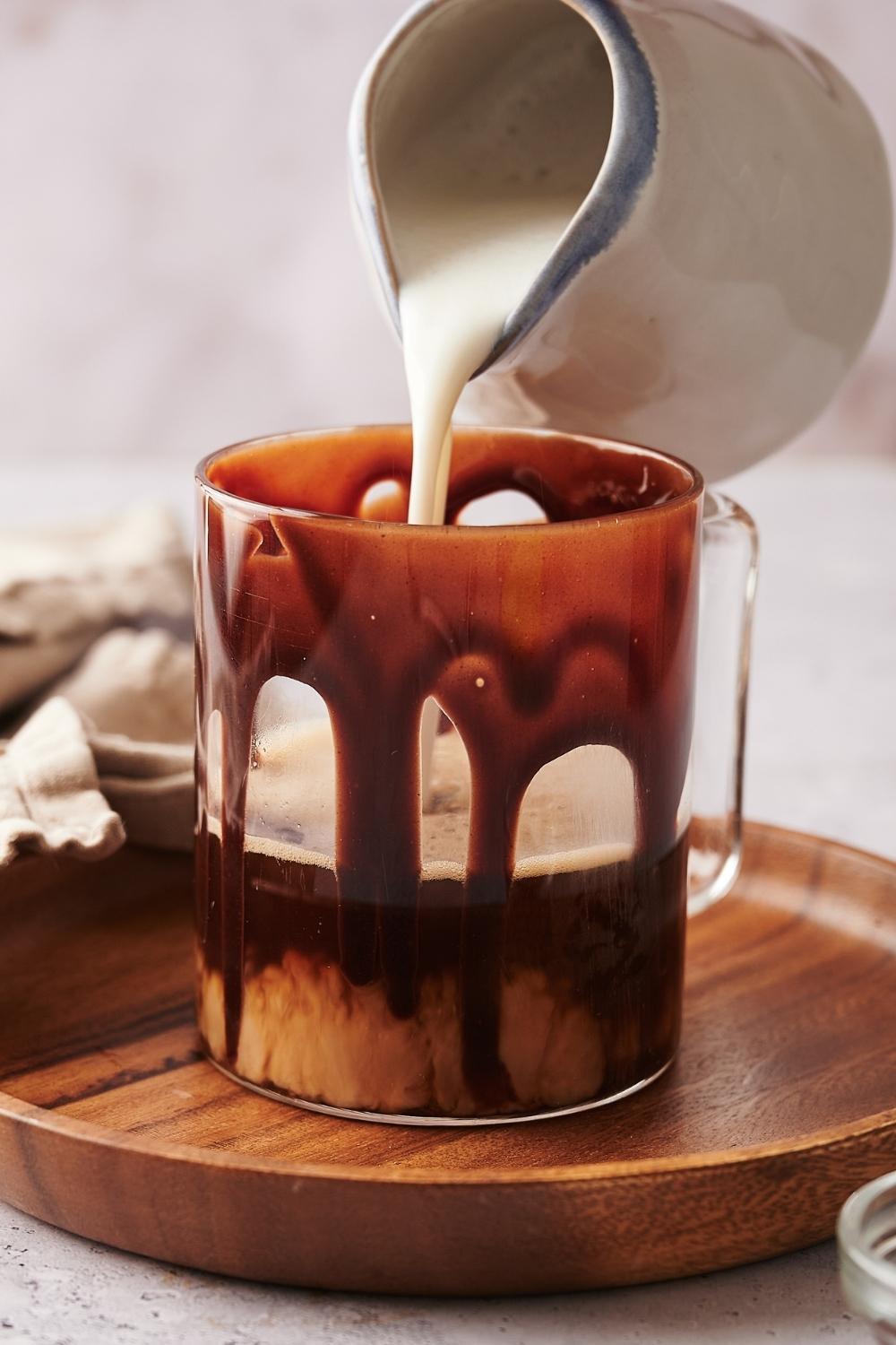 A clear glass mug with chocolate syrup drizzled on the sides. Steamed milk is being poured into the mug.