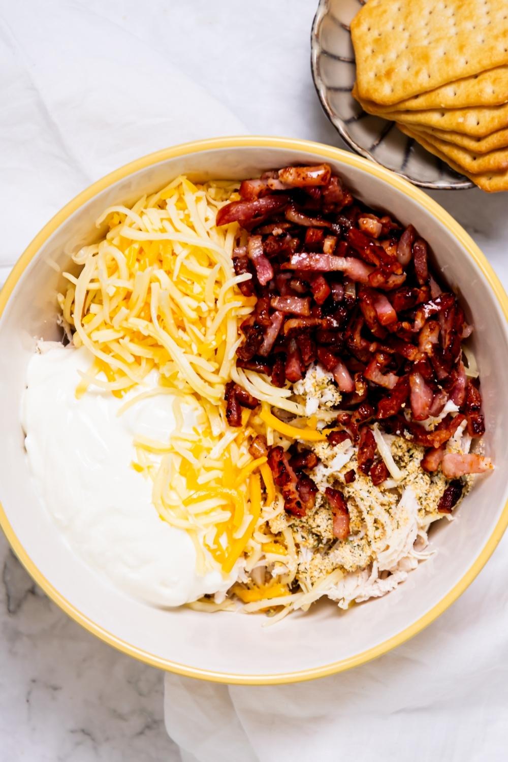 A mixing bowl with crispy bacon, shredded chicken, shredded cheese, and sour cream.
