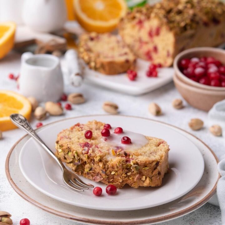 A slice of cranberry bread on a white plate with the whole loaf of cranberry bread behind it.