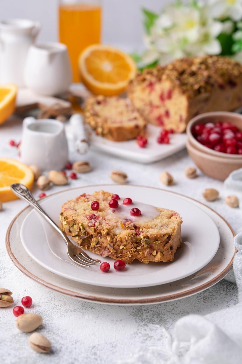 A slice of cranberry bread on a white plate with the whole loaf of cranberry bread behind it.