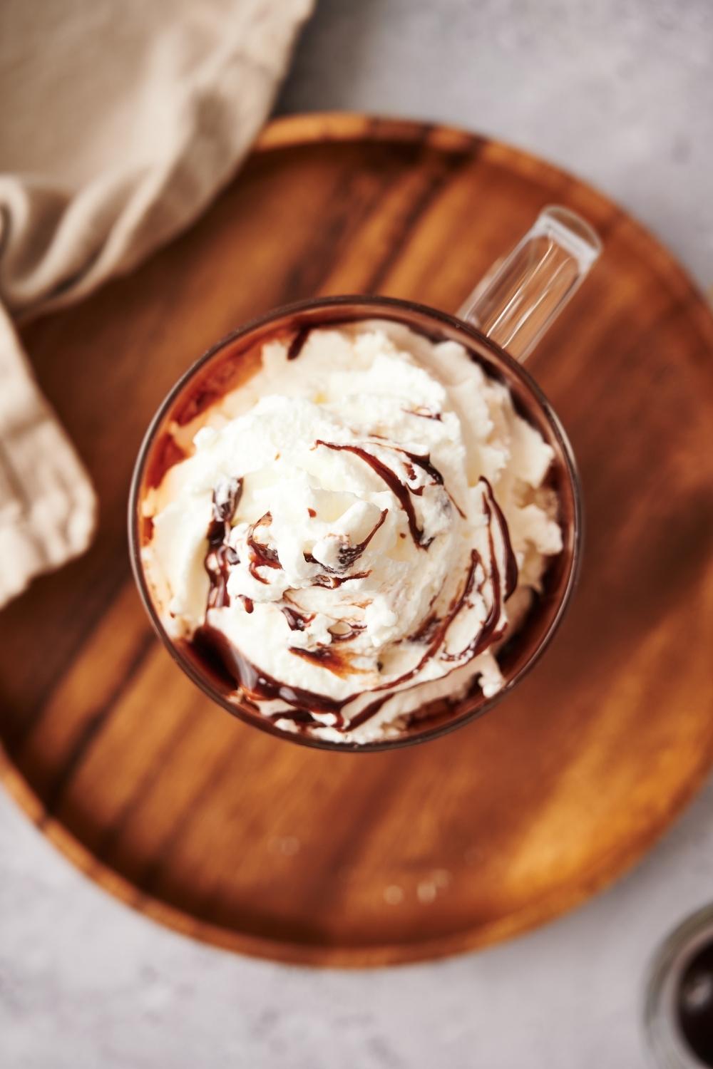 Whipped cream with chocolate sauce drizzled on top in a glass mug on a wooden plate.