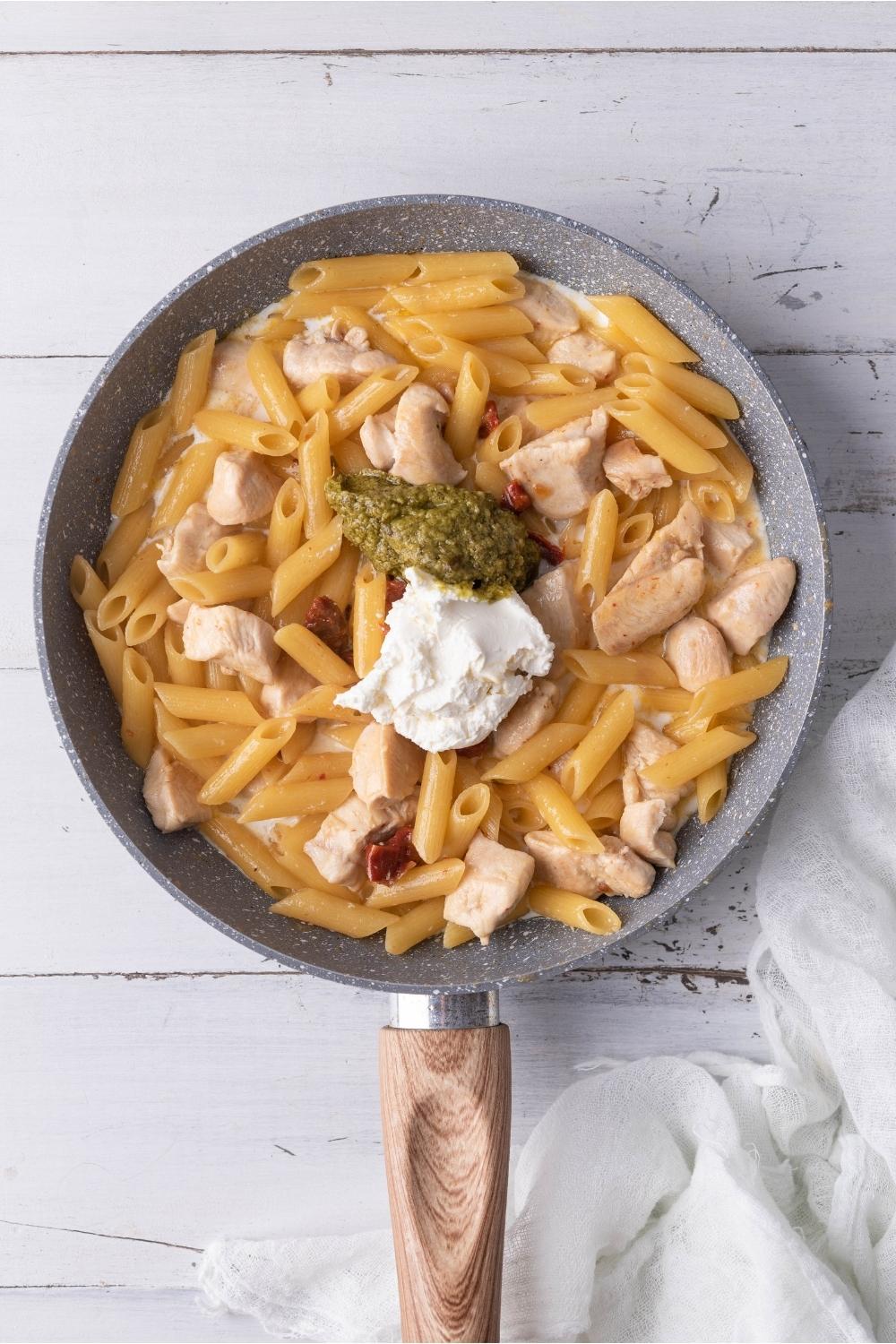 Skillet with pesto chicken pasta ingredients, pesto and cream cheese freshly added.