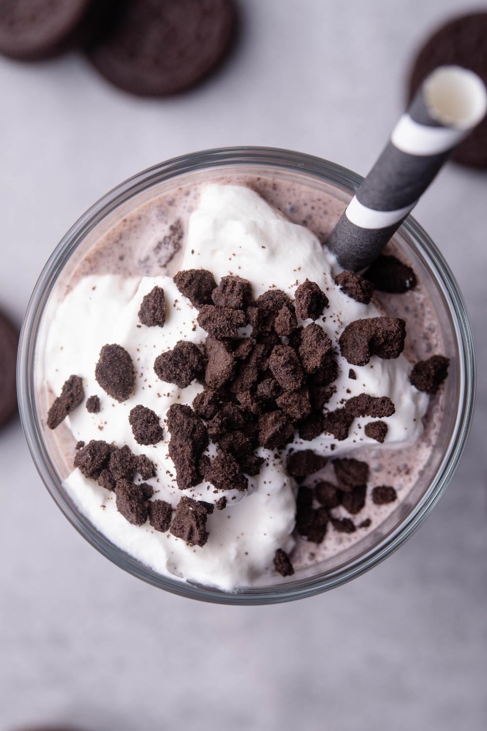 An overhead view of a tall glass with homemade oreo mcflurry in it, topped with whipped cream and oreo cookie crumbles. A striped paper straw is in the cup.