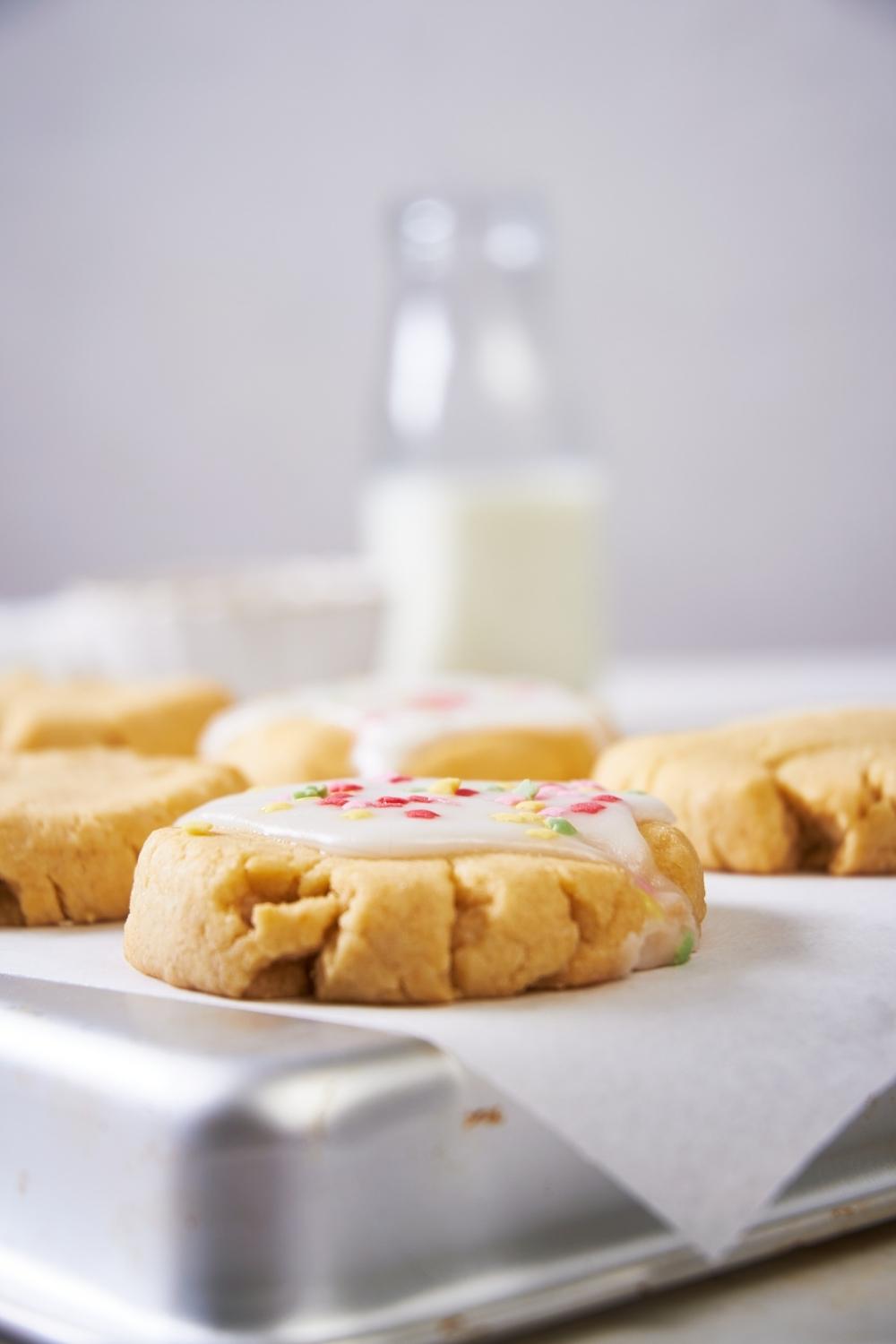 close up shot of a glazed sugar cookie on parchment paper over an upside down baking tray. a glass bottle of milk sits in the back