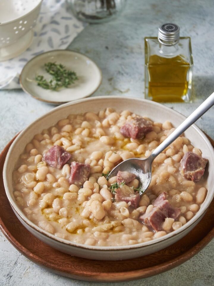 A big bowl of great northern beans with ham served on a wooden plate tray. A spoon is in it.