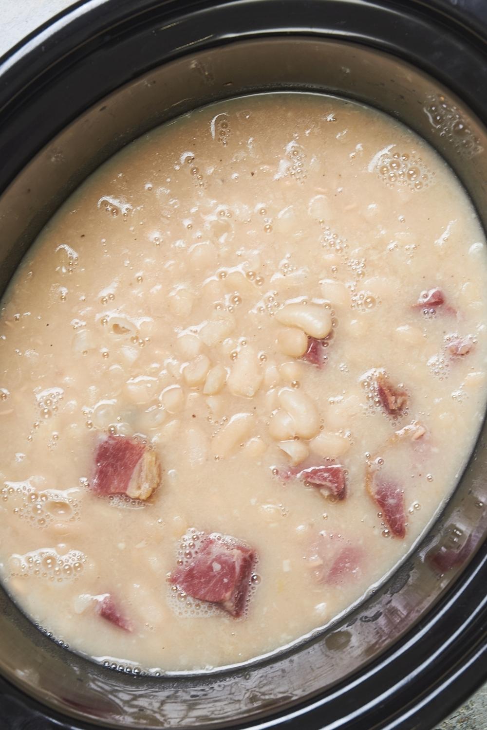 A crock pot with cooked great northern beans with ham in it.