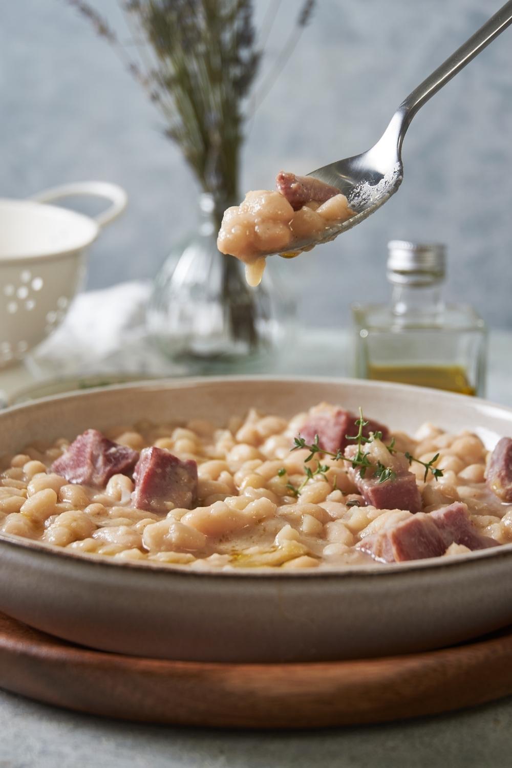 A big bowl of great northern beans with ham served on a wooden plate tray. A spoon has a scoop of it held above the plate.
