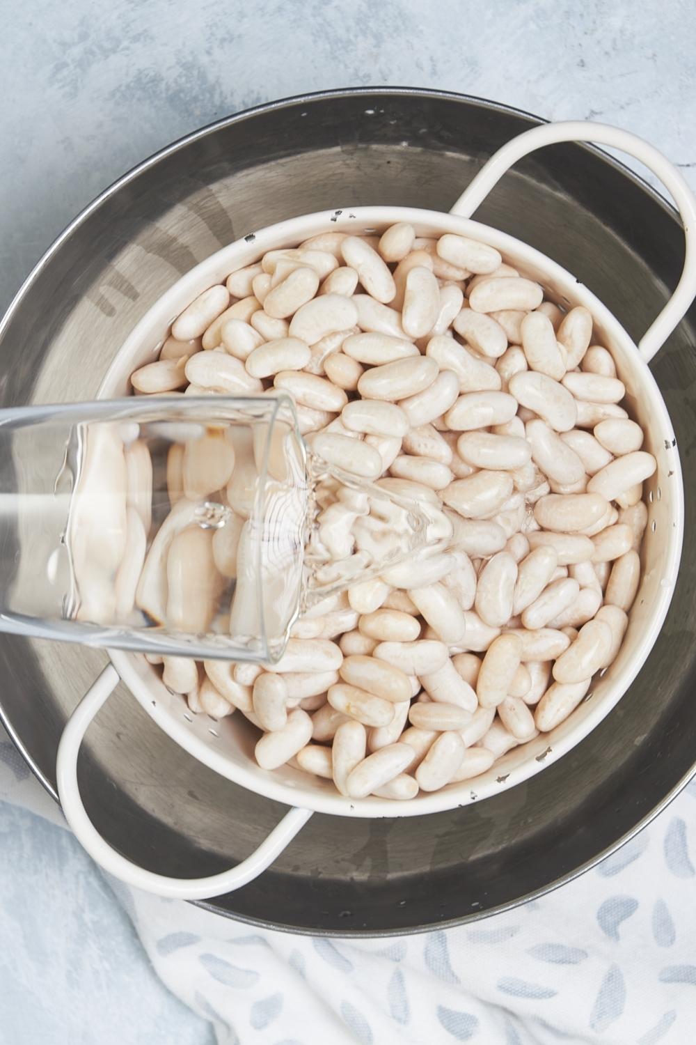 A bowl with a strainer full of dry great northern beans. A pitcher of water is filling the bowl to soak the beans overnight.