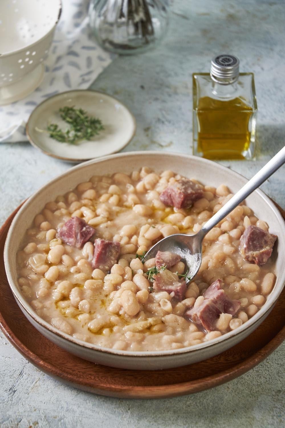 A big bowl of great northern beans with ham served on a wooden plate tray. A spoon is in it.