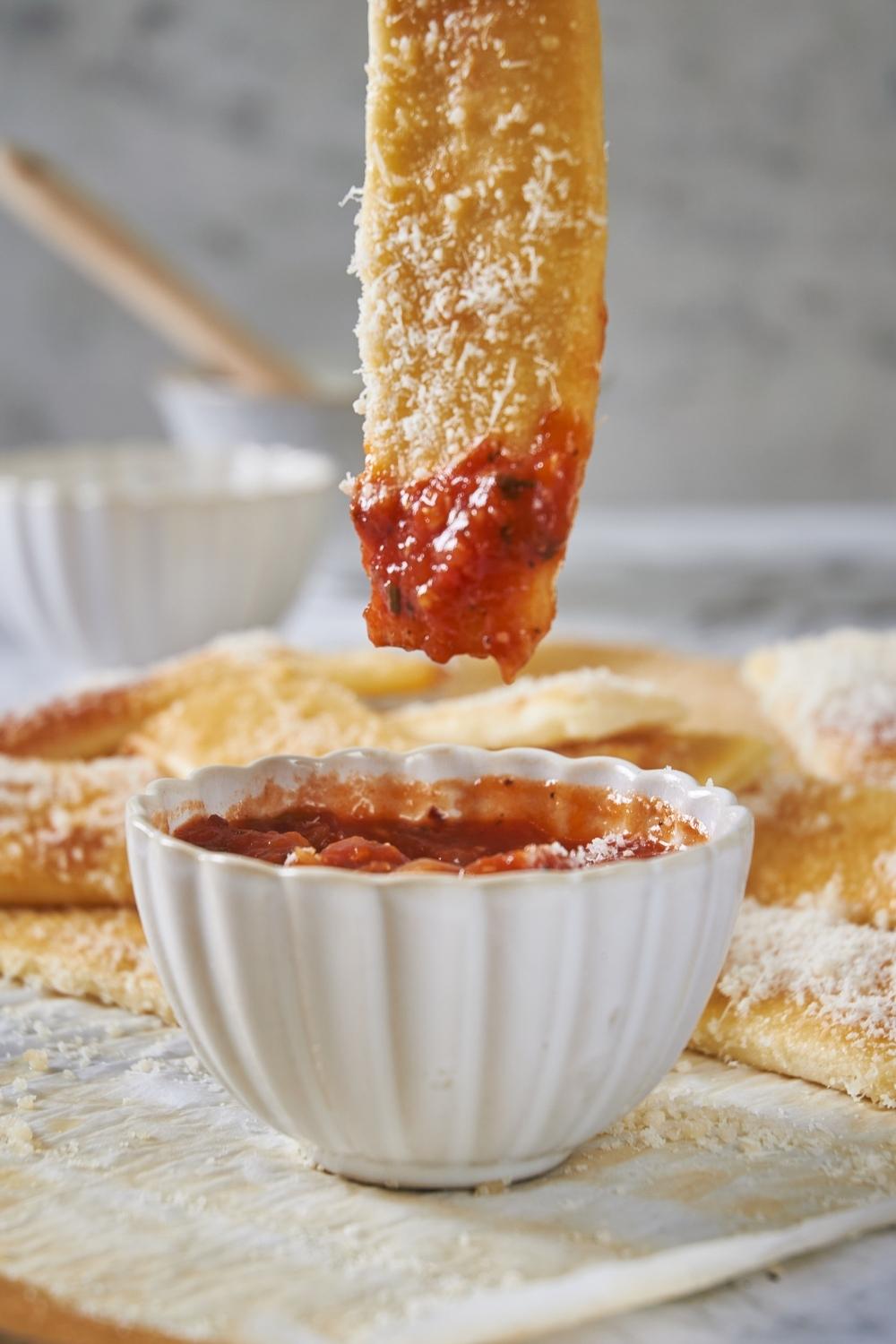 A stick of little caesars crazy bread has been dunked into the side bowl of pizza sauce and is getting lifted out. The remaining little caesars crazy bread sticks are on a cutting board in the background.