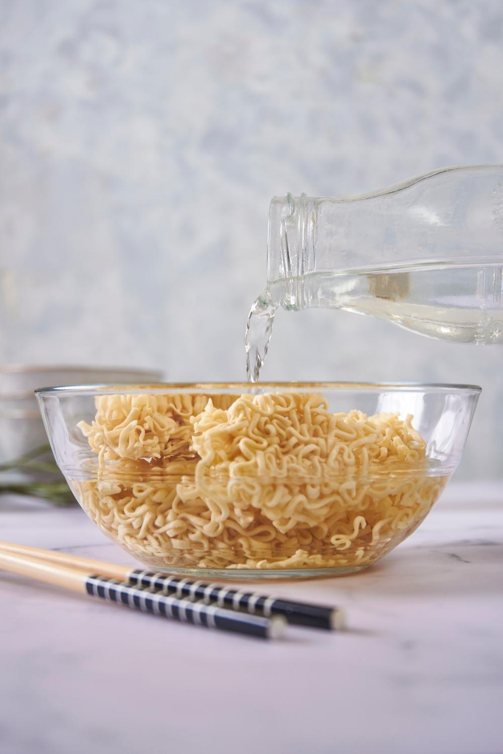 A clear bowl of uncooked ramen with a pitcher pouring water into the bowl and chopsticks next to the bowl.