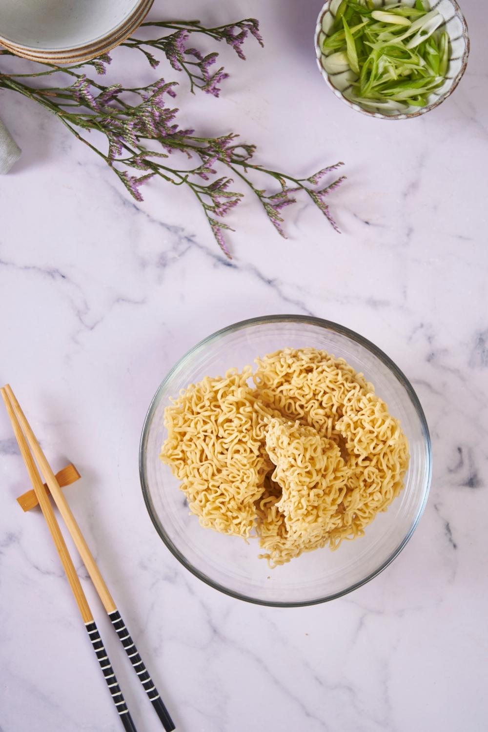 A clear bowl of uncooked ramen noodles next to a pair of chopsticks and a small bowl of diced green onion.