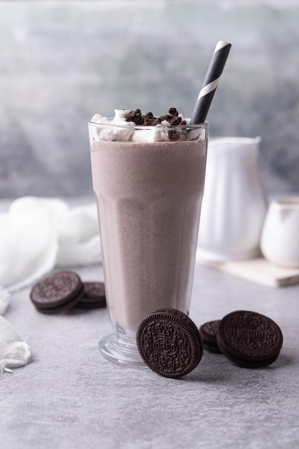 A tall glass with homemade oreo mcflurry in it, topped with whipped cream and oreo cookie crumbles. A striped paper straw is in the cup. Oreo cookies sit around the base of it on the counter. And 2 pitchers sit on a wooden cutting board in the background.