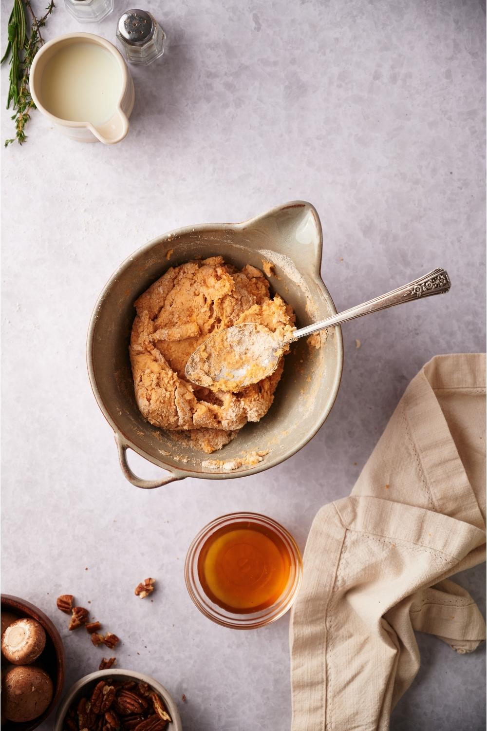 A large mixing bowl of biscuit dough with a spoon in the bowl next to a small bowl of maple syrup.