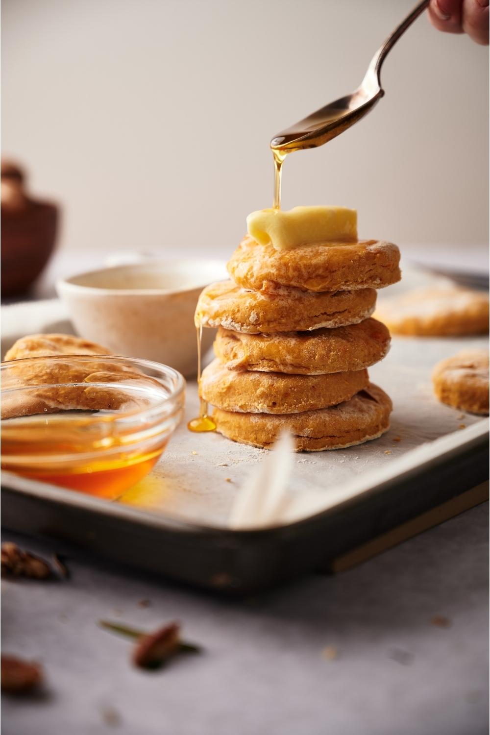 A stack of sweet potato biscuits on a baking sheet with a hand pouring maple syrup over top of the biscuits.