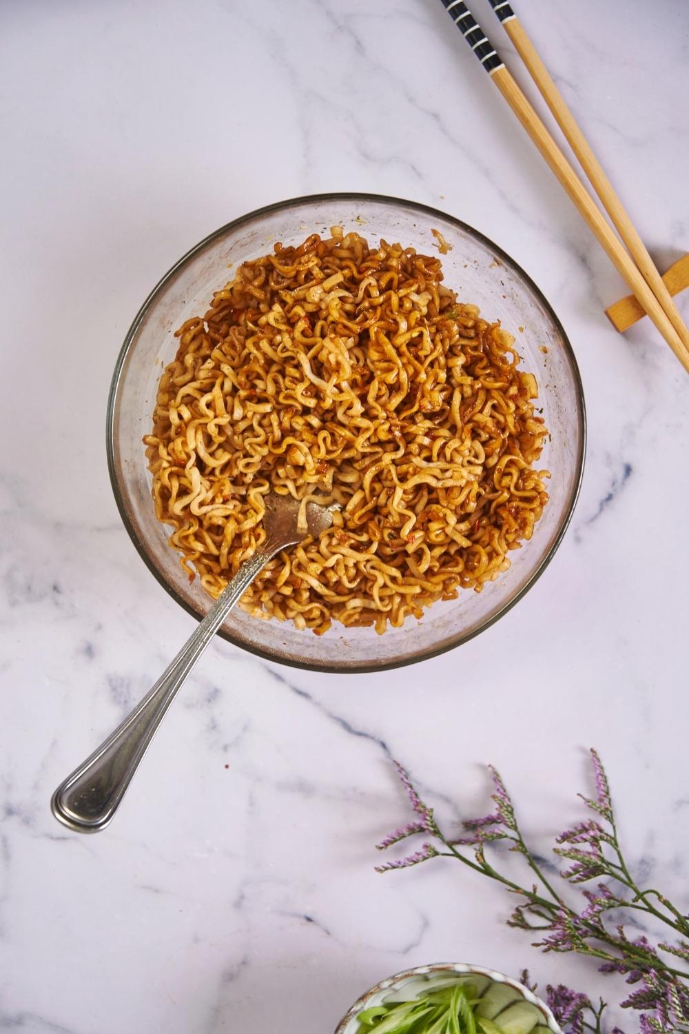 Microwaved ramen in a clear bowl with a fork in the bowl and chopsticks next to the bowl.