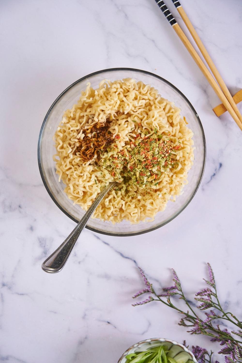 Cooked ramen in a clear bowl with a seasoning packet added and not yet mixed in, with a fork in the bowl and chopsticks next to the bowl.