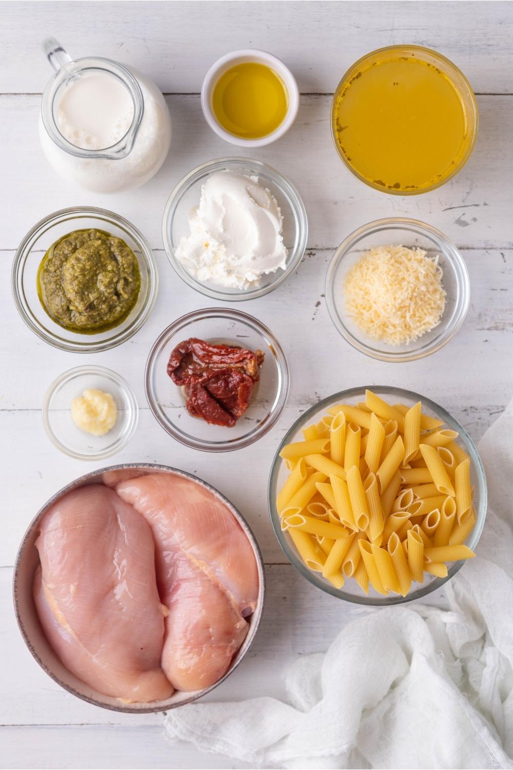 An assortment of ingredients to make pesto chicken pasta including raw chicken, penne pasta, chicken broth, cream cheese, pesto, and seasonings.