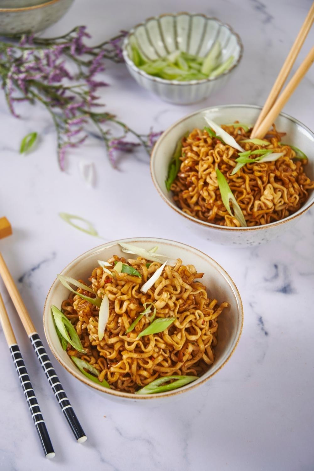 Two bowls of microwaved ramen, one with chopsticks in the bowl and one with chopsticks next to the bowl, both garnished with green onion.