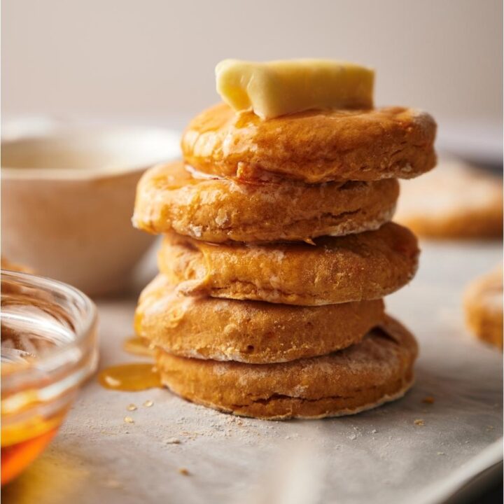 A stack of sweet potato biscuits on a baking sheet lined with parchment paper, the biscuits are covered in maple syrup and a pad of butter is melting on top of the stack of biscuits.