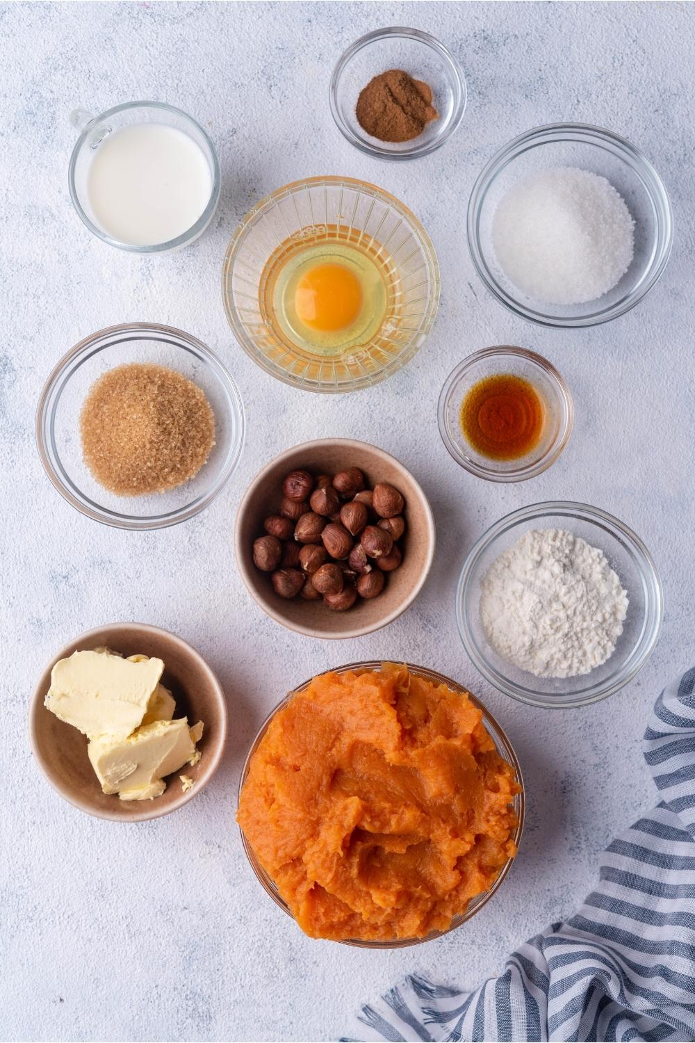 An assortment of ingredients for sweet potato souffle including bowls of sweet potato puree, butter, hazelnuts, egg, sugar, flour, and spices.