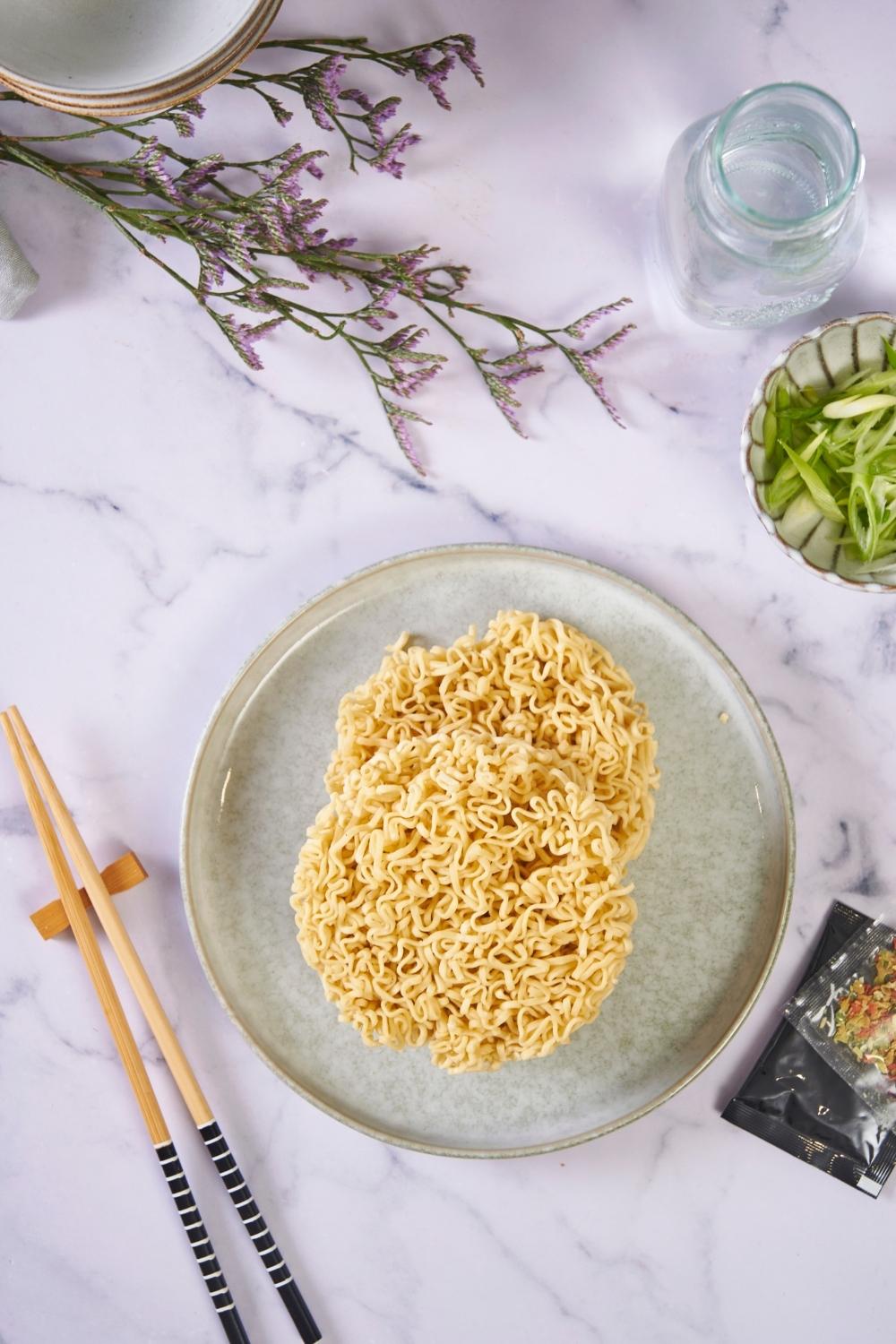 Two discs of uncooked ramen noodles on a blue plate, next to a pair of chopsticks and a small bowl of diced green onion.