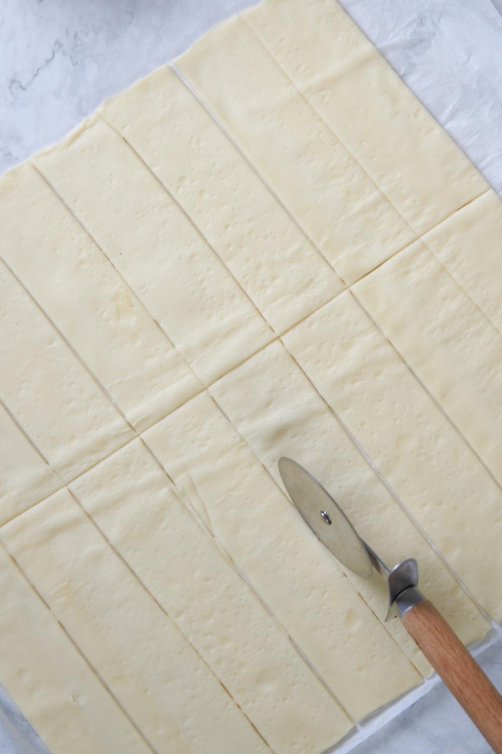 A parchment paper with pizza dough on it. A pizza cutter is cutting the dough into strips.