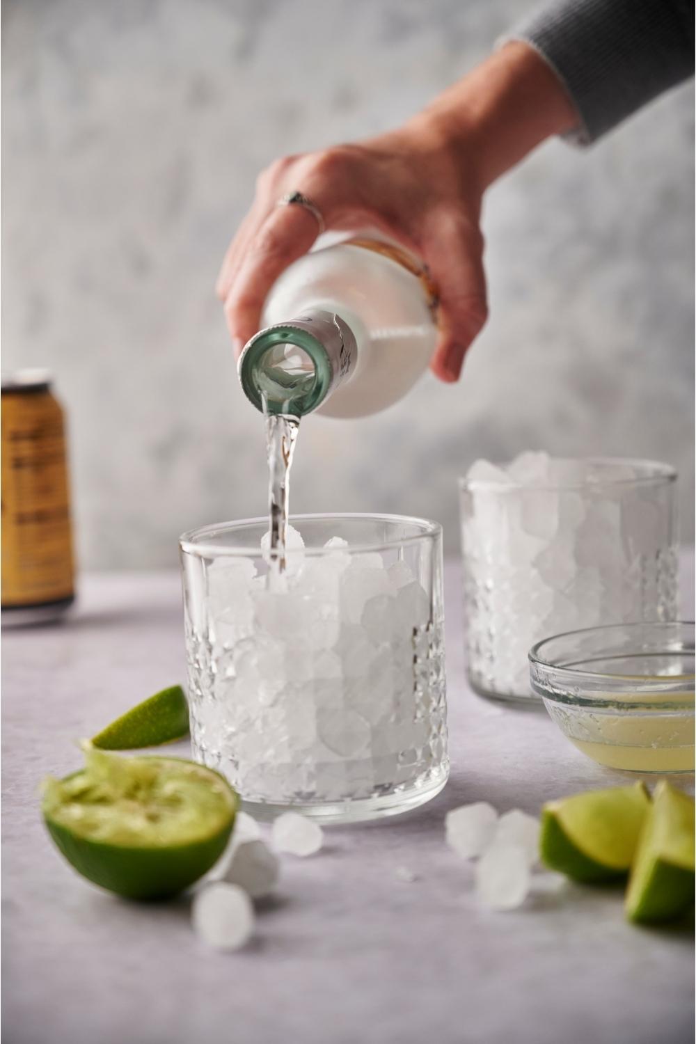 A hand pouring tequila from a bottle into a glass of ice that is surrounded by juiced lime halves and ice cubes.