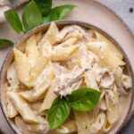 Shredded chicken on top of penne pasta in a creamy sauce in a bowl on a plate.