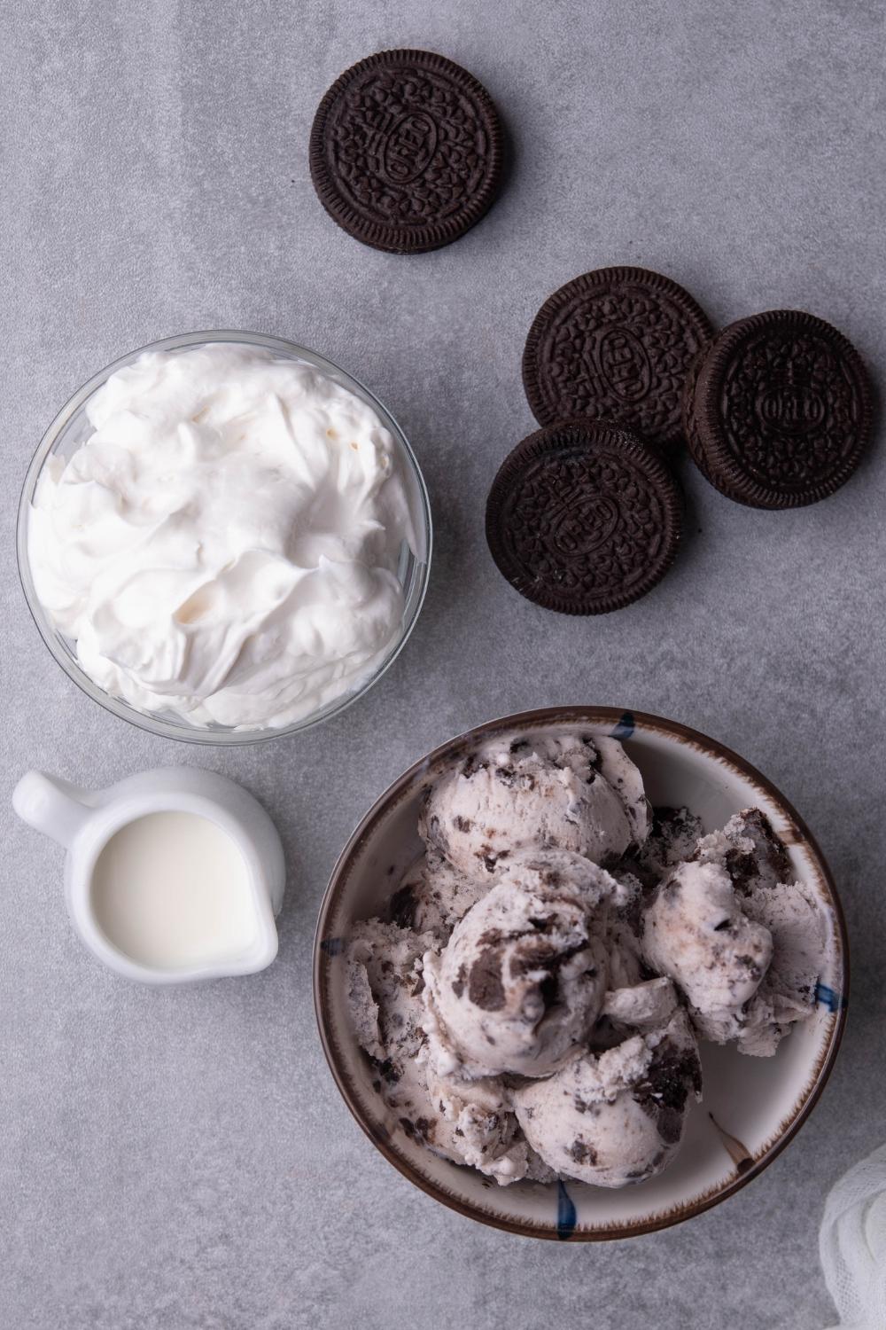 A countertop with a bowl of cookies and cream ice cream, a bowl of cool whip, a small pitcher with milk, and 4 whole oreo cookies.
