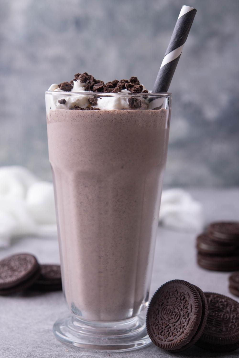 A tall glass with homemade oreo mcflurry in it, topped with whipped cream and oreo cookie crumbles. A striped paper straw is in the cup. Oreo cookies sit around the base of it on the counter.
