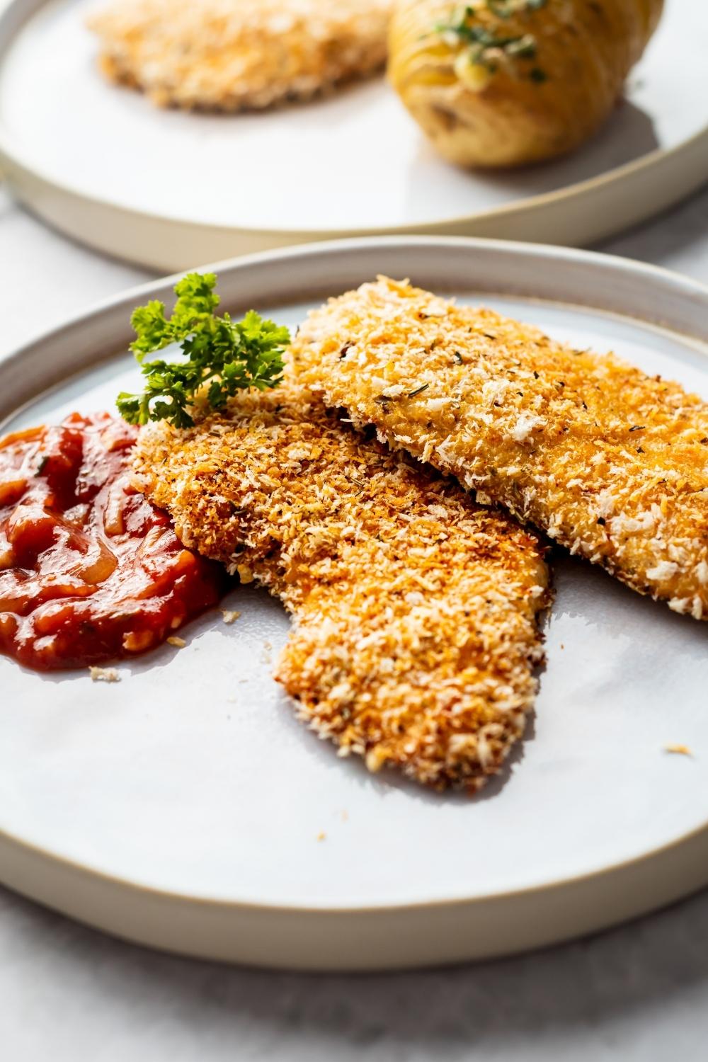 two pieces of panko breaded chicken on a white plate with a sprig of parsley and some marinara sauce