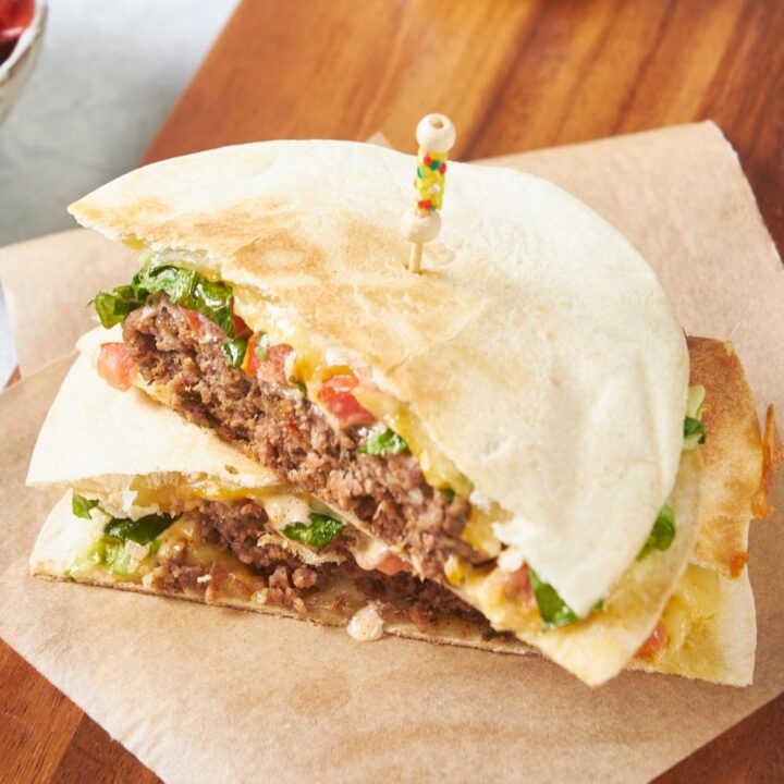 Half of a quesadilla burger on top of another half of a quesadilla burger on top of piece of parchment paper on a wooden board.