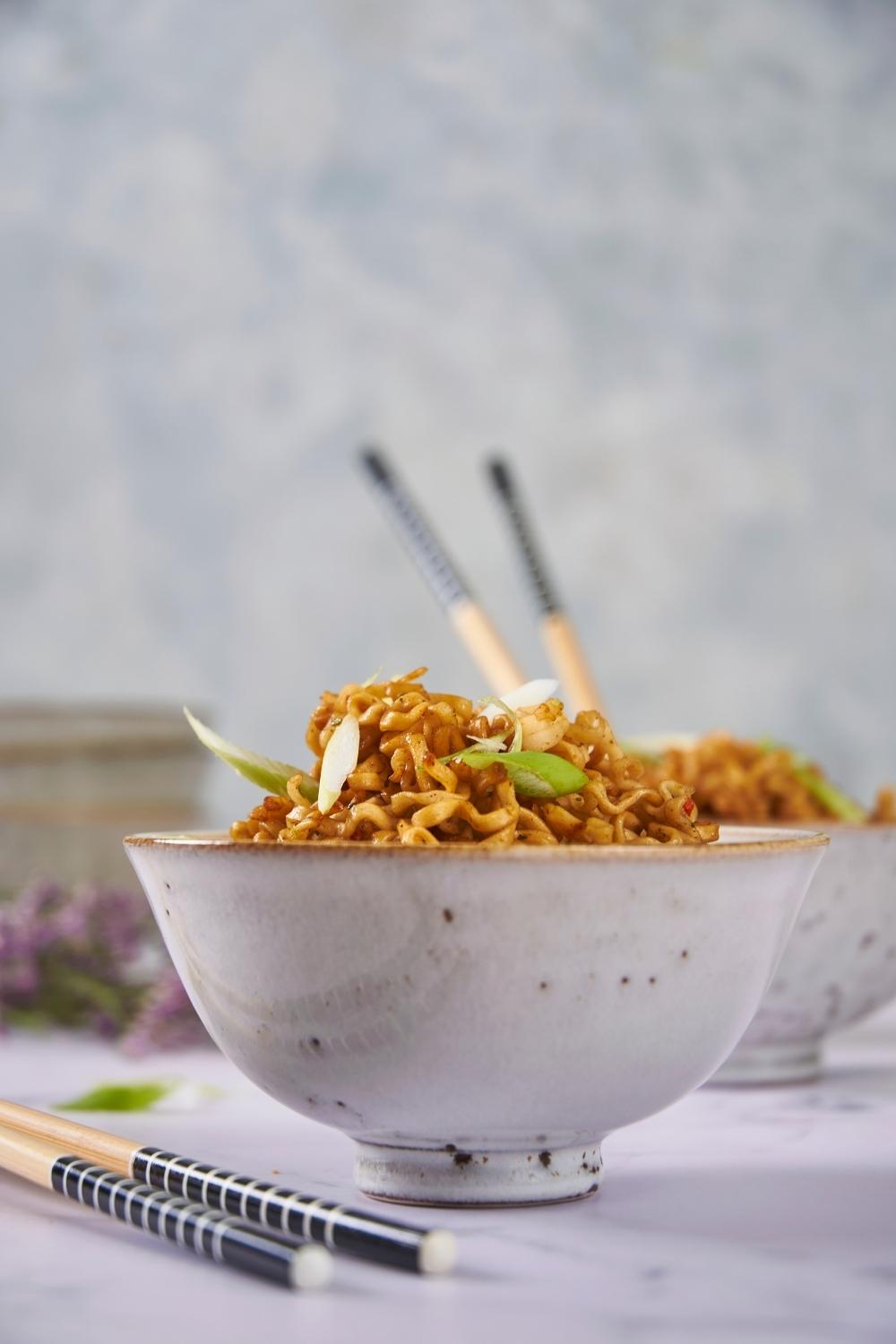 Microwaved ramen in a white bowl with chopsticks next to the bowl and a second bowl of ramen in the background.