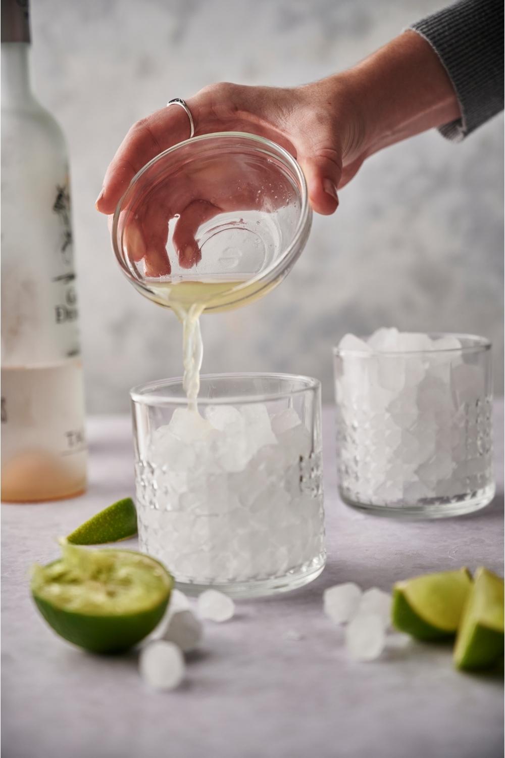 A hand pouring fresh lime juice into a glass of ice next to ice cubes and juiced lime halves.