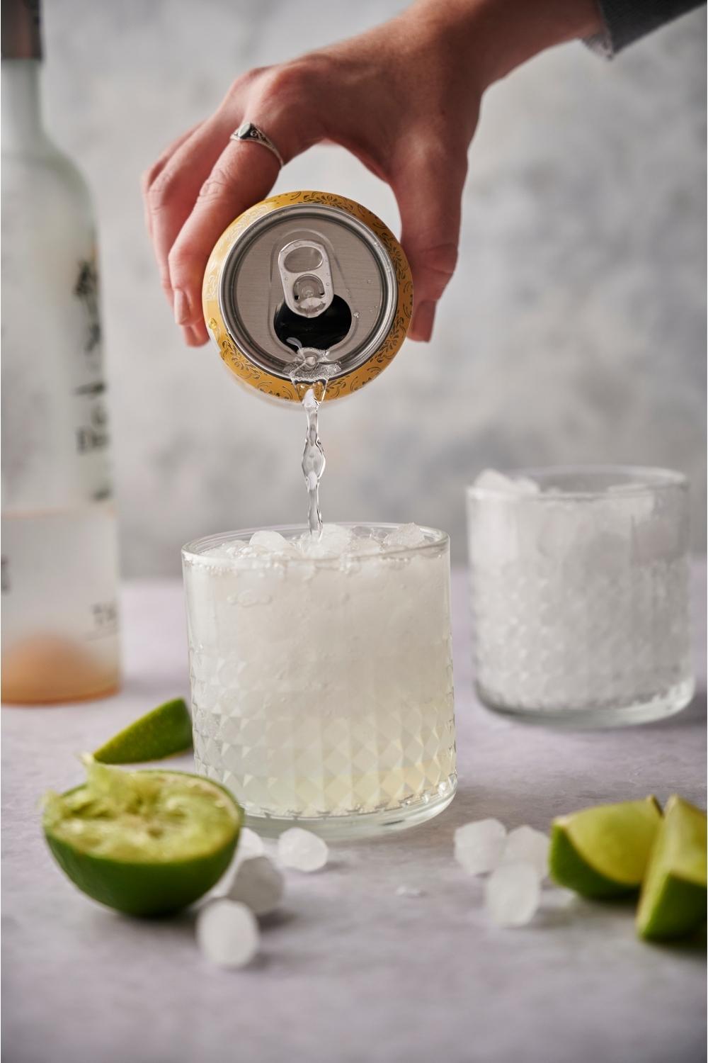 A hand pouring a can of sparkling mineral water into a cocktail surrounded by ice cubes and juiced limes.