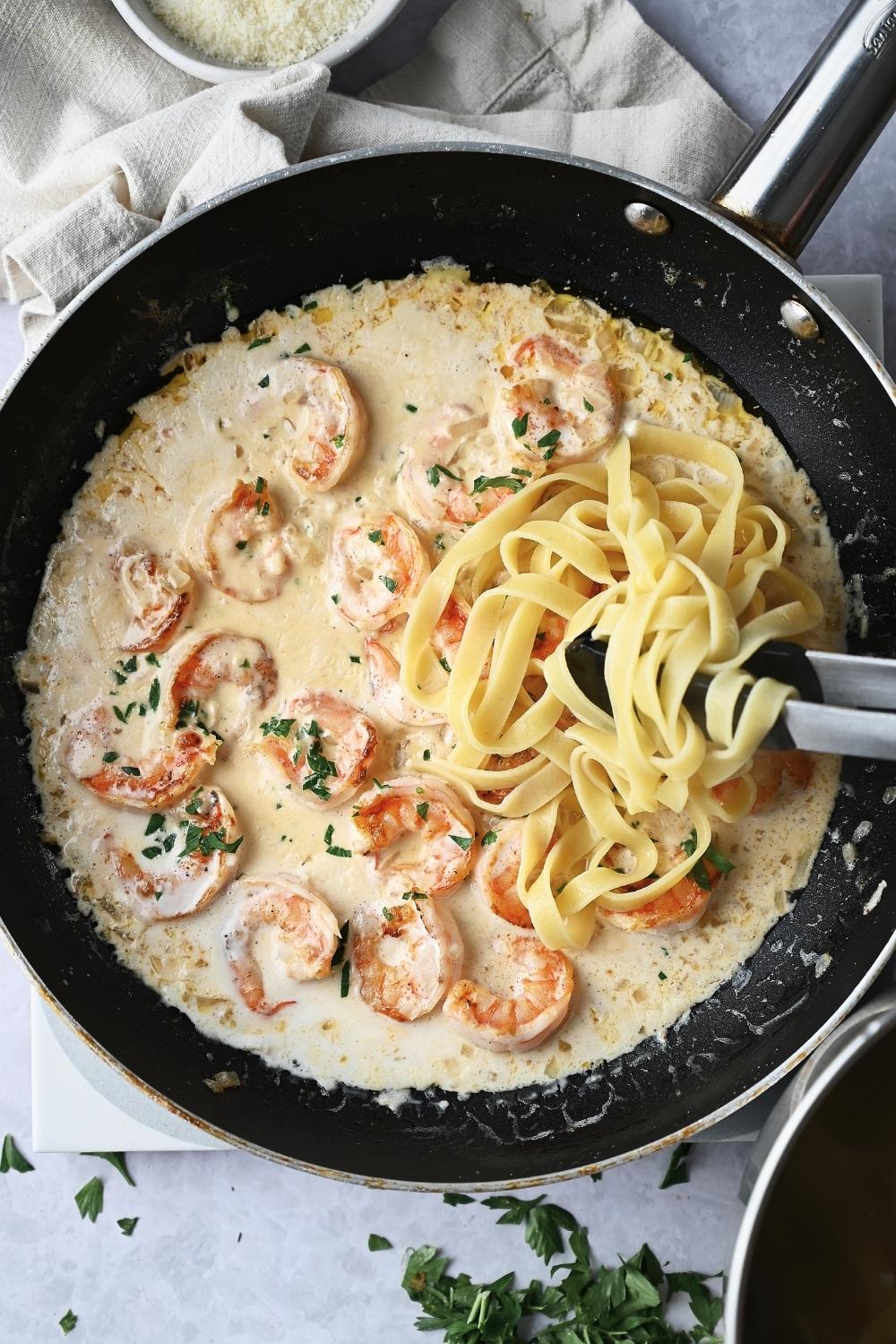 A pan with onion, garlic, and shrimp with garlic cream sauce reducing. Cooked Fettuccini noodles are being added.