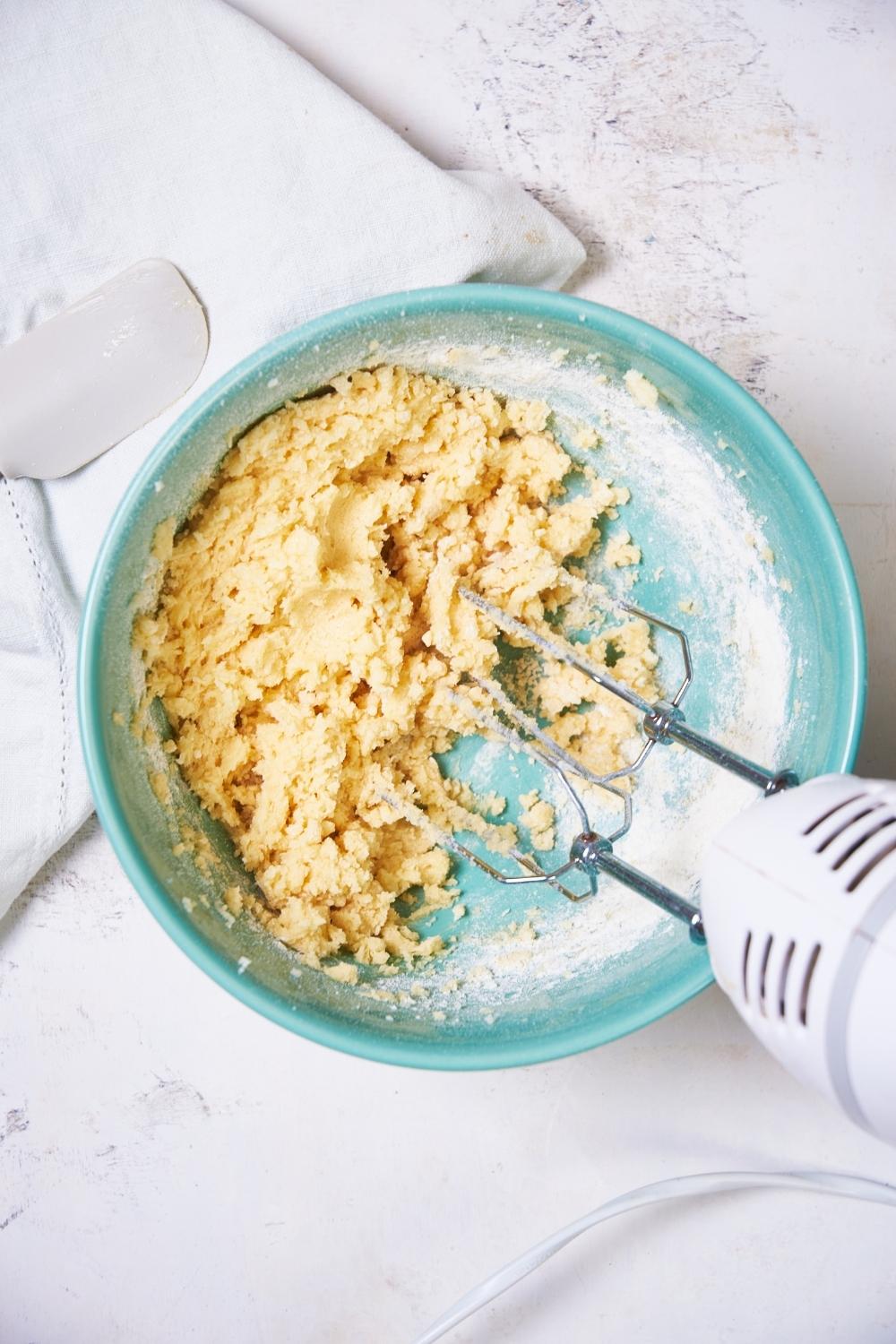sugar cookie dough in a teal bowl being mixed together with an electric mixer. a tea towel and a rubber spatula sit to the side