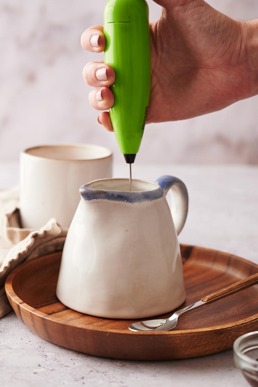 A small pitcher with warmed milk in it, a hand is holding a frother in the pitcher.