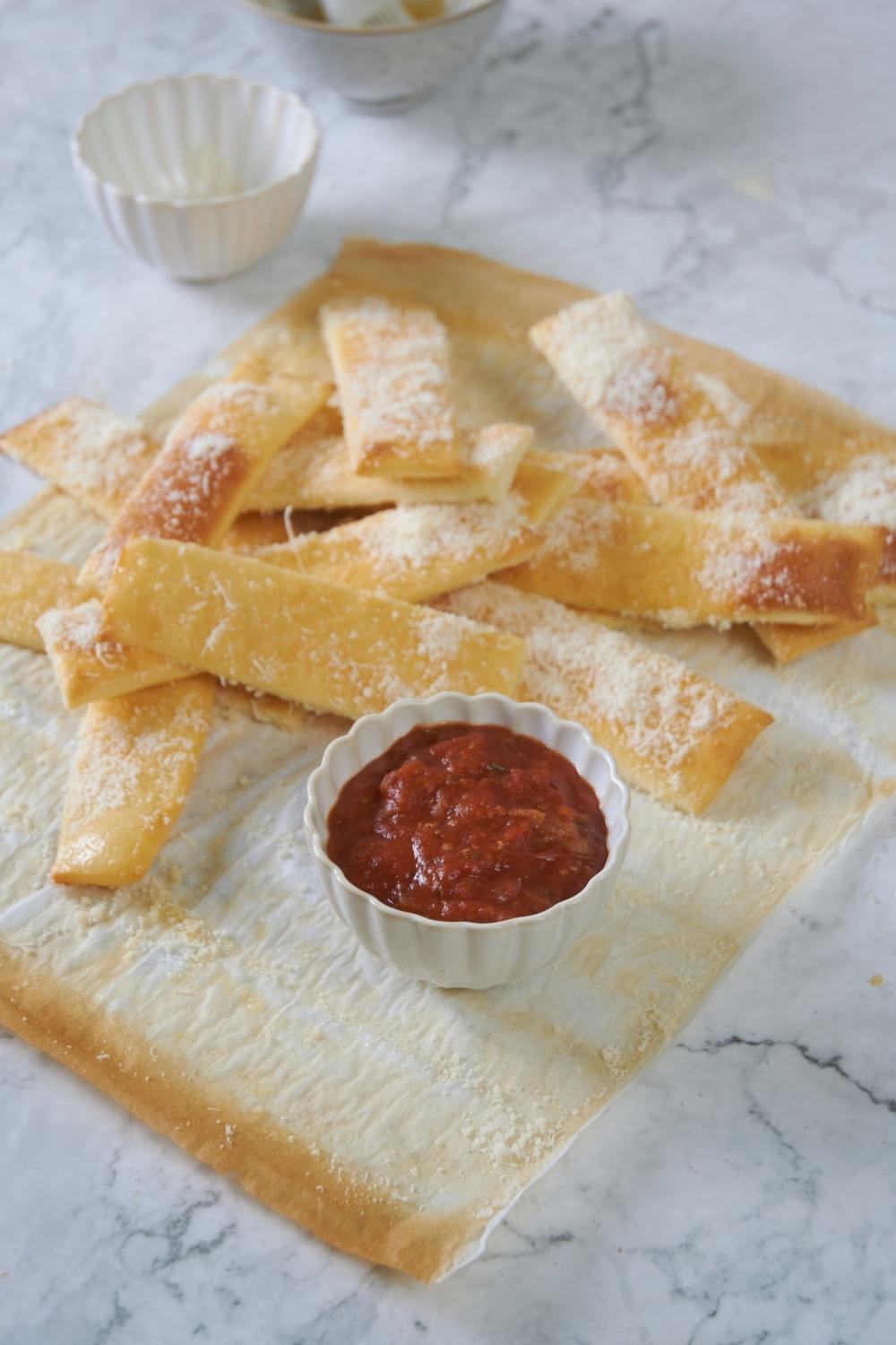 A pile of homemade little caesar's crazy bread sticks lay on a parchment paper sheet. A side bowl with pizza sauce sits next to them.
