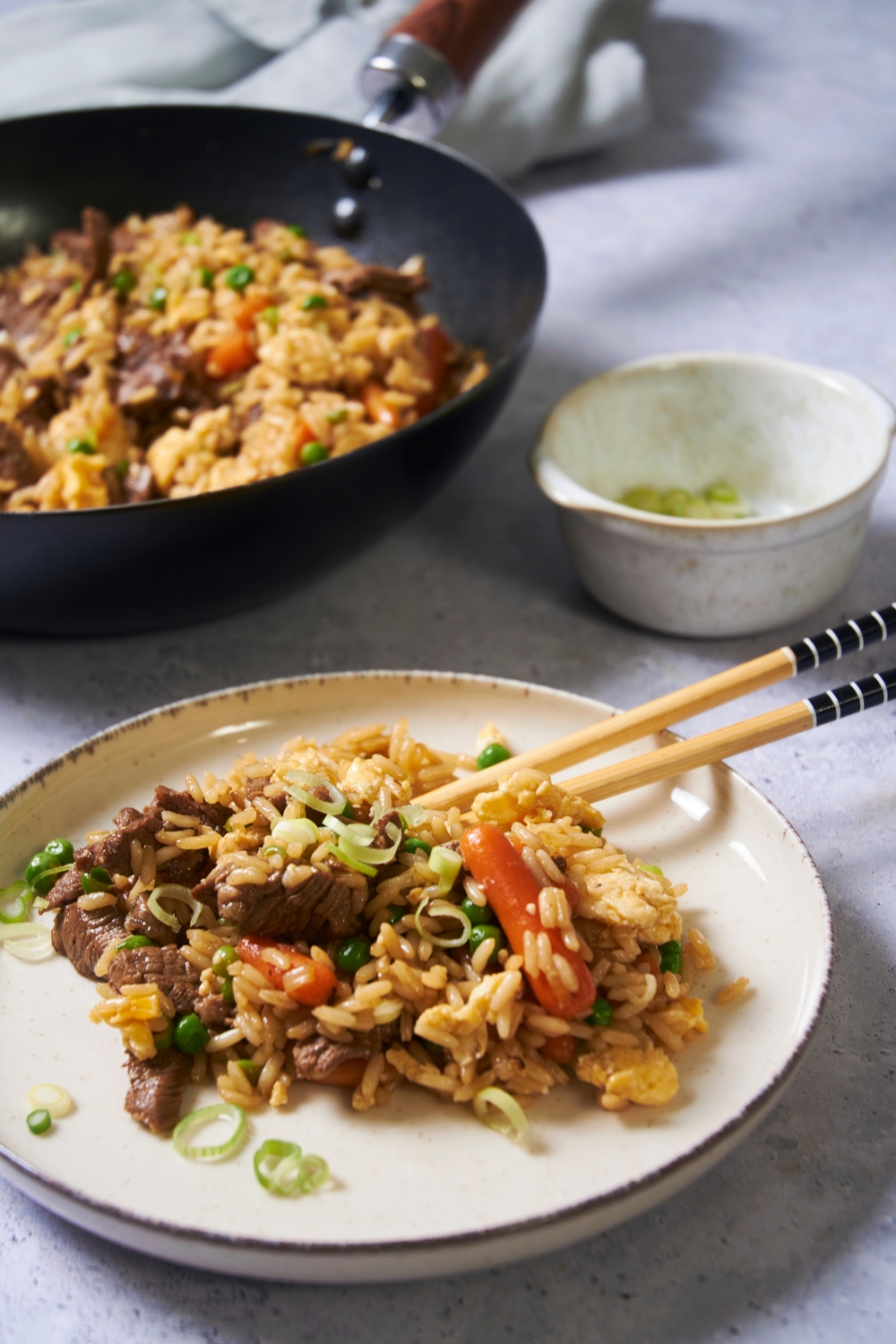 A white plate with beef fried rice and chopsticks on the plate. Behind the plate there is a small bowl of scallions and a black skillet filled with fried rice.