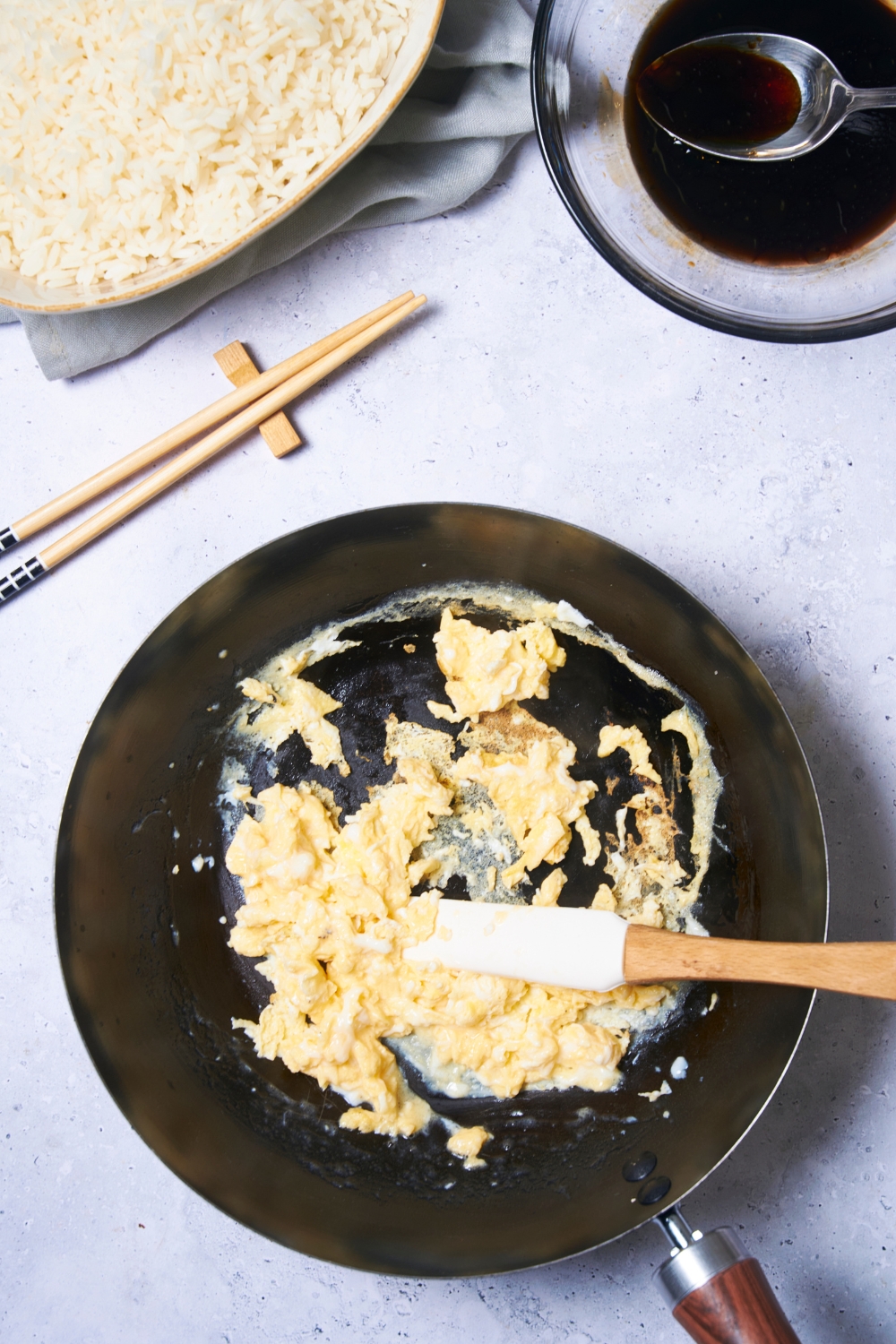 A black skillet with scrambled eggs and a plastic spatula in the skillet. Next to the skillet is a pair of chopsticks, a bowl of white rice and a bowl of a brown sauce mixture with a spoon in the bowl.