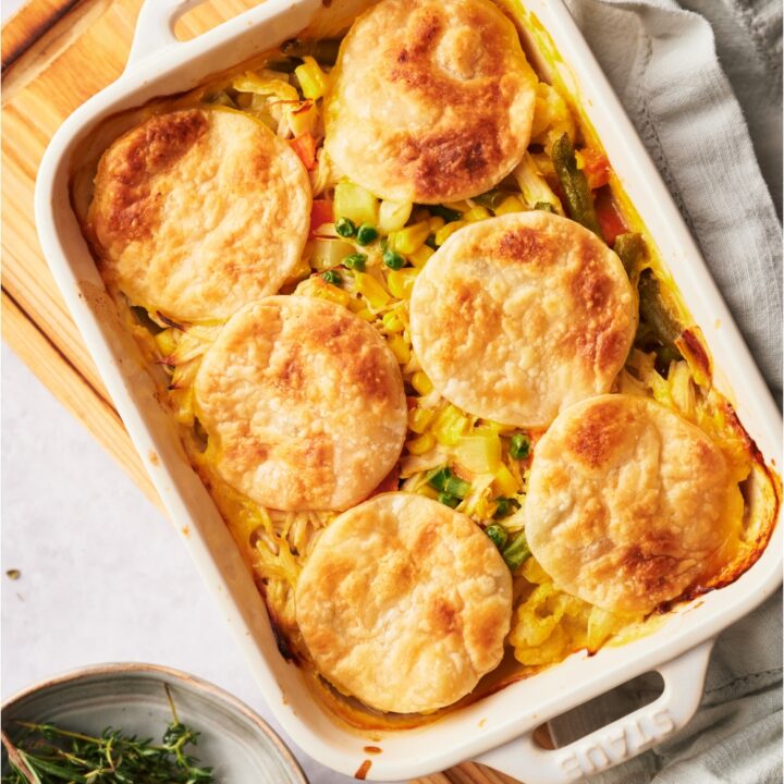 A white casserole dish with chicken pot pie casserole freshly baked. The dish is on a wood board and next to it there is a bowl of fresh rosemary and a dish towel.