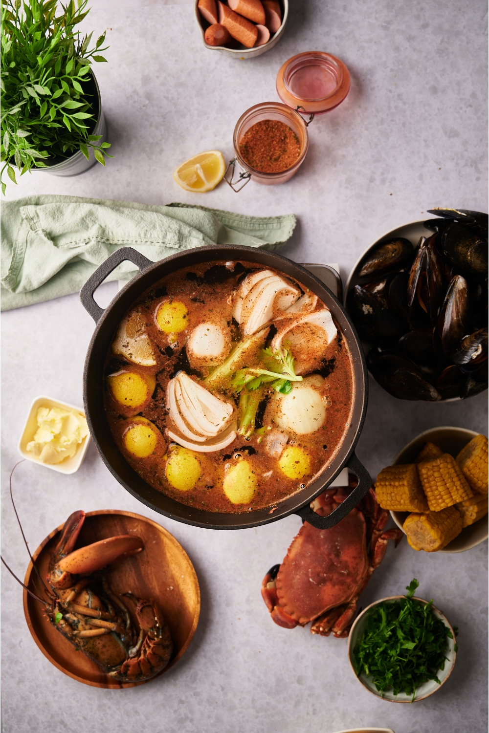 Black Dutch oven filled with onions, celery, potatoes and lemon wedges in a brown broth. Surrounding the Dutch oven is an assortment of ingredients including lobster, crab, clams, sausages, corn, fresh herbs, and spices.